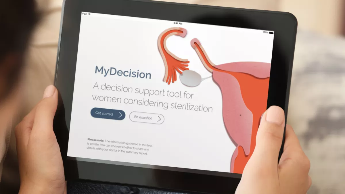 A person holds a tablet displaying the MyDecision app, a decision support tool for women considering sterilization, featuring an anatomical illustration of the uterus and options to get started in English or Spanish.